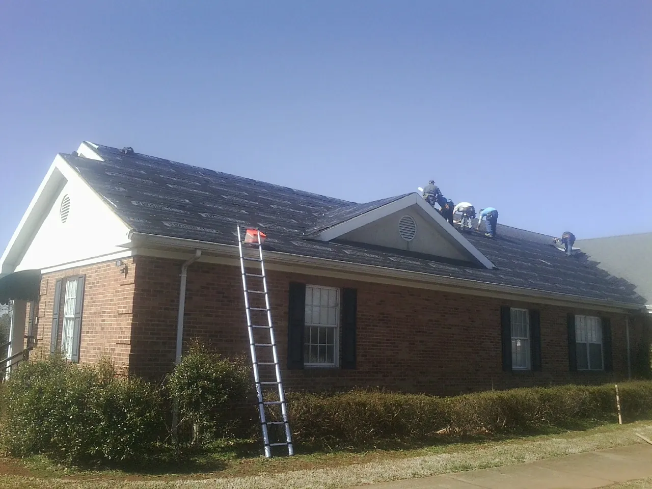 Crew tearing shingles off a brick ranch roof with a ladder set — Metal Roof Repair work by our crew in Spofford