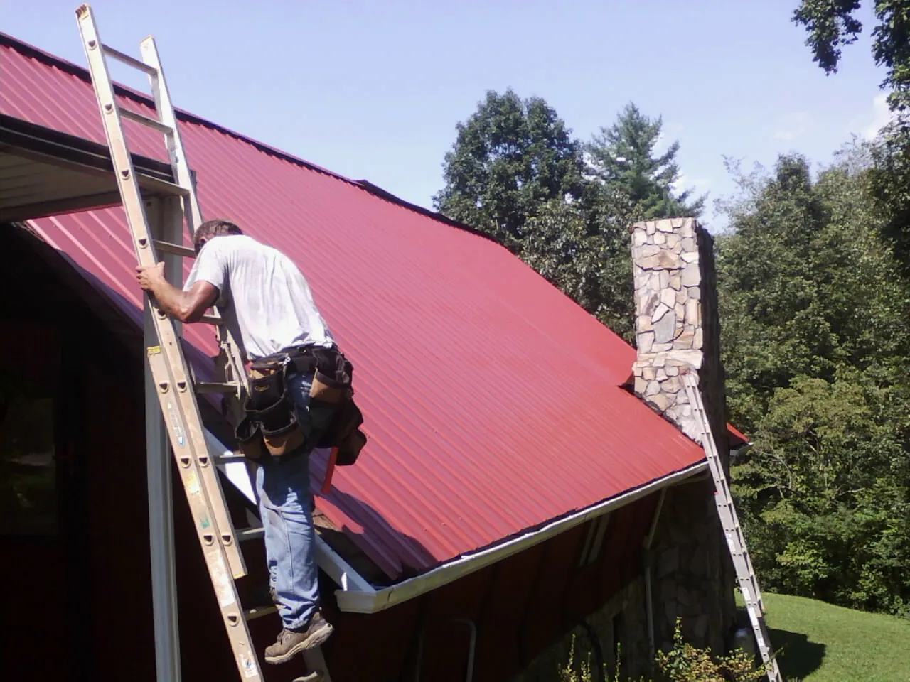 Roofer on a ladder beside a red metal roof and stone chimney — Soffit & Fascia Repair by Roofing Brothers in Spofford